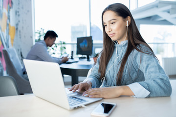 Positive attractive young lady in earphones sitting at table and analyzing information on laptop while working in contemporary open space office
