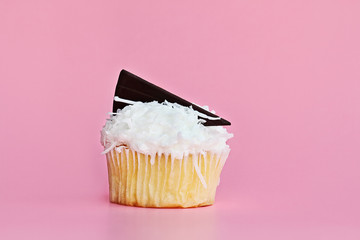 Pretty coconut frosted cupcake decorated with a wedge of chocolate against a pink background.