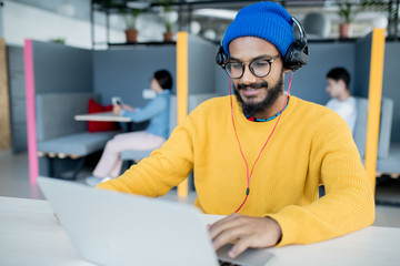 Positive satisfied middle-eastern app developer in headphones sitting at table and using laptop...