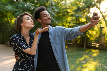 Smiling young multiethnic loving couple standing posing in park outdoors hugging take a selfie by phone.