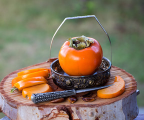 Kakifrucht in einem kleinen Schälchen mit Fruchtscheiben und Obstmesser auf einer Eukalyptus-Holzplatte