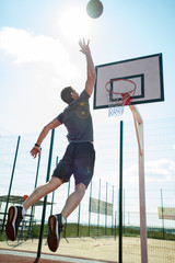 Full length back view portrait of young sportsman shooting slam dunk in basketball court outdoors,...