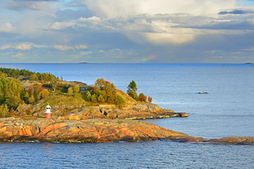 Rocky island with lighthouse of Helsinki archipelago at sunset. Distant rainbow in sea. Finland