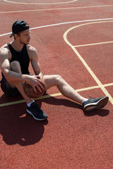 Full length portrait of handsome muscular man sitting on floor in outdoor basketball court and looking away pensively