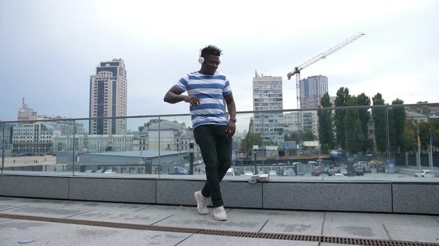 Handsome trendy african american hipster in white headphones walking along street and moving in afrohouse style dance over cityscape background. Cheerful black man dancing outdoors. Slo mo.