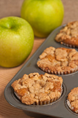 Top view, close distance of four, freshly baked, homemade apple crumb muffins cooling in teflon, metal, cup cake baking sheet and two green apples