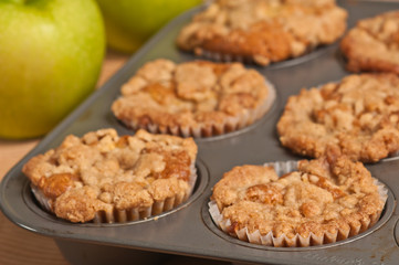 Top view, close up of six, freshly baked, homemade apple crumb muffins cooling in teflon, metal, cup cake baking sheet and two green apples