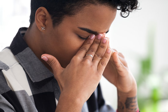 Close Up Young African Stressed Businesswoman Sitting Indoors Alone Suffering From Headache Feels Unwell Touch Nasal Root Closing Eyes. Frustrated Black Female Employee Have A Problem In Work Concept