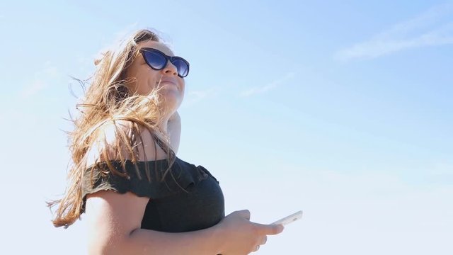 Happy woman in glasses uses a smartphone, on the beach in windy weather, in slow motion. Medium Shot