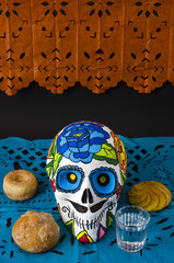 Styrofoam skull with flowers and mustache in a day of the dead offering altar with orange and blue cut paper, bread and mezcal