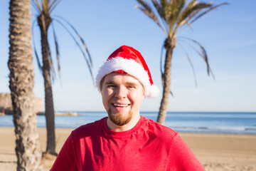 Obraz premium Christmas, people, holidays concept - Young man wearing a Santa hat smiling at the beach