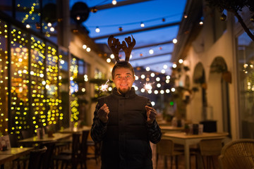 Fototapeta premium People, holidays and christmas concept - surprised man in deer's horns holding two bengals light or sparklers outdoors