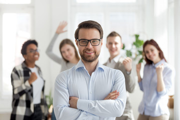 Smiling millennial confident businessman standing with arms crossed looking at camera with happy cheerful business partners team at background. Successful teamwork, business owner company boss concept