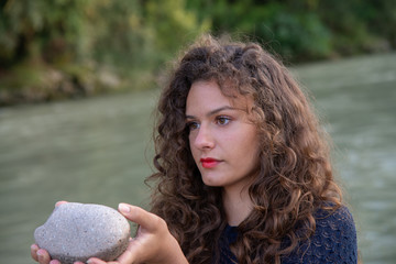 Girl with long wavy brown hair. With a stone in his hands along the bank of a river. Warm sun at sunset. Fine white sand The girl is wearing a perforated blue dress