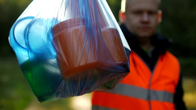 A man in a signal orange vest holds a package with garbage on the background of nature, forest, close-up, garbage collection, a janitor, volunteer