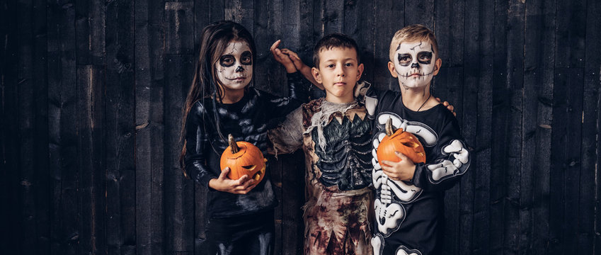 Three Multiracial Kids In Scary Costumes Posing With Pumpkins In An Old House.