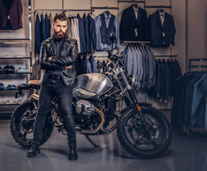 Fototapeta na wymiar Brutal male dressed in black jacket posing with crossed arms near retro sports motorbike at the men's clothing store.