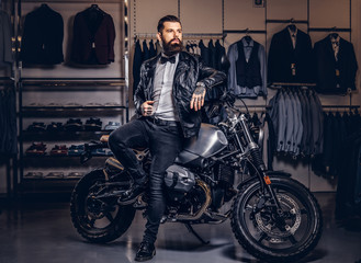 Obraz na płótnie Canvas Stylish tattooed bearded man with dressed in black leather jacket and bow tie posing near retro sports motorbike at men's clothing store.