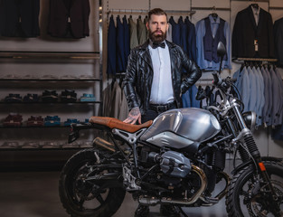 Fototapeta na wymiar Stylish tattooed bearded man with dressed in black leather jacket and bow tie posing near retro sports motorbike at men's clothing store.