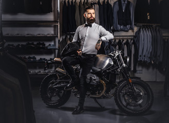 Fototapeta na wymiar Elegantly dressed tattooed male with stylish beard holds black leather jacket posing near retro sports motorbike at the men's clothing store.
