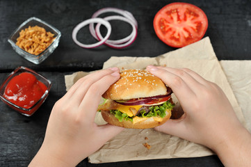 Cheeseburger in children's hands. On the table are paper, onion rings, cups with ketchup and dried onions, a tomato. Black background. Close-up. View from above. 