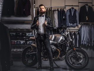 Plakat Stylish tattooed bearded man with dressed in black leather jacket and bow tie posing near retro sports motorbike at men's clothing store.