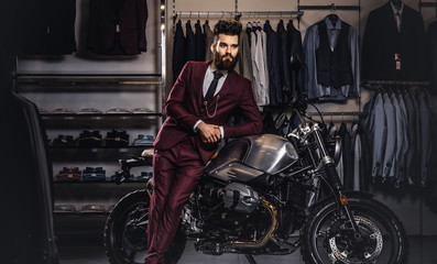 Fototapeta na wymiar Handsome man with a stylish beard and hair dressed in vintage red suit posing near retro sports motorbike at men's clothing store.