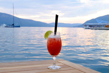 A Glass of Cocktail with lemon on wood table  with seascape and skyline in the evening with sunset tone style   Sea wave background Background of stunning turquoise sea Travel concept Red cocktail