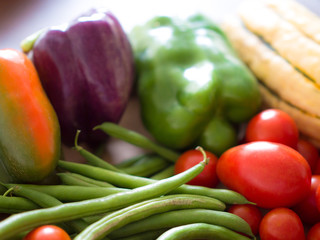 Close up photograph of several types of fresh organic vegetables including green beans, green pepper, purple pepper, and orange sweet pepper, cherry tomatoes, and squash.