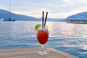 A Glass of Cocktail with lemon on wood table  with seascape and skyline in the evening with sunset tone style   Sea wave background Background of stunning turquoise sea Travel concept Red cocktail