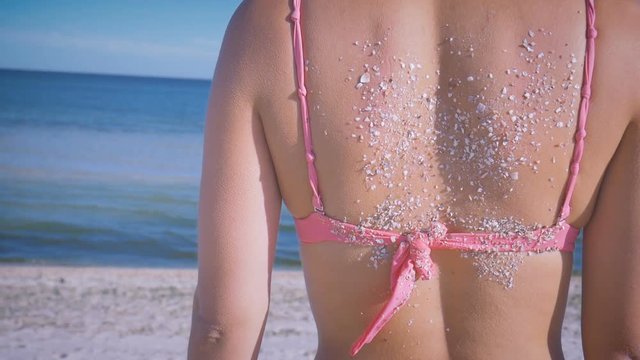 Close up swarthy back of a girl in a pink swimsuit in the sand and shell shards on the background of the sea