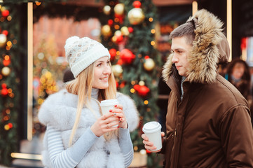 christmas portrait of happy couple with hot mulled wine or tea walking on city streets decorated for holidays, christmas trees with lights and toys on background