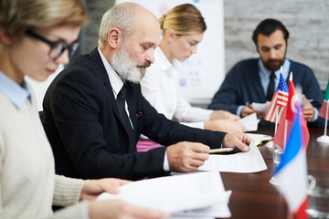 Senior bearded businessman or politician reading document among his young colleagues at conference