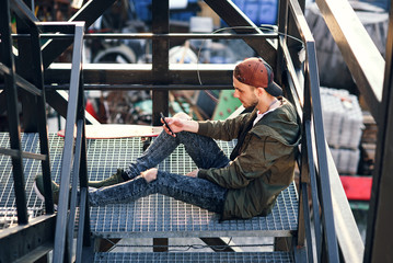 Young handsome hipster man with skateboard using smartphone on the iron stairs of the industrial building.
