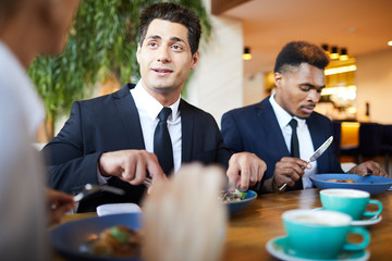 Positive handsome young businessman in formal suit sitting at table and eating salad while chatting with colleagues at dinner in restaurant