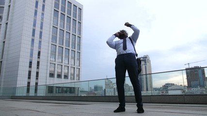 Joyful afriacan american businessman celebrating promotion with his fist clenched after phone call from head office over modern building background. Successful executive in victory gesture outdoors.