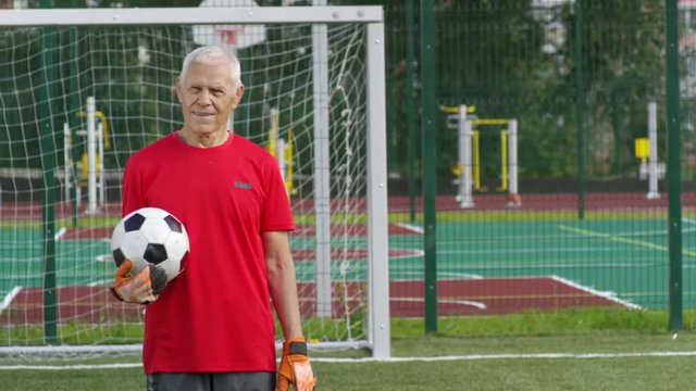 Portrait of active Caucasian elderly man in t-shirt and goalkeeper gloves standing at goal net and holding football while posing for camera, tracking shot