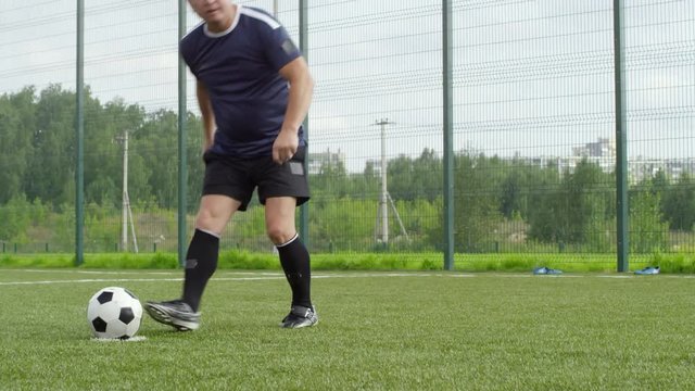 Mature Asian man in sports clothing putting football on ground and kicking it while training and playing outdoors on summer day