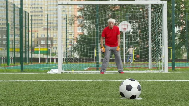 Tracking shot of active senior man in sports clothing and goalkeeper gloves standing at goal net, catching football and throwing it back towards camera while playing with friends