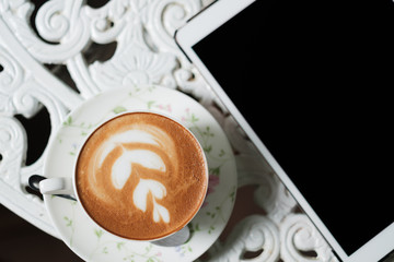 Cup of latte art coffee and tablet computer on white vintage desk.