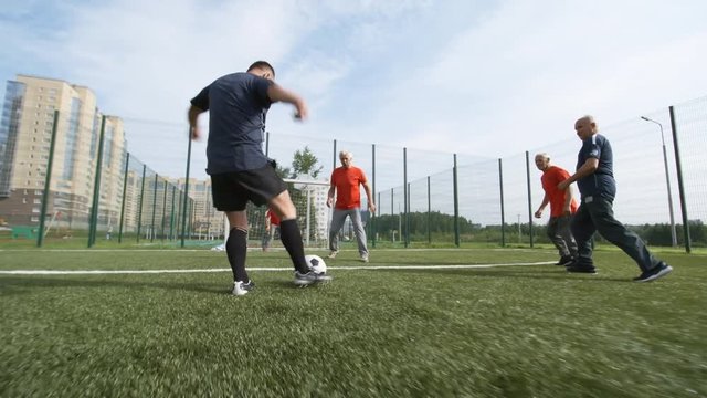 Group of five elderly male friends playing football outdoors on warm summer day, one of them kicking ball and scoring goal for his team