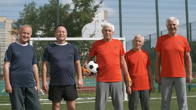 Group of five active retired men in red and blue t-shirts standing in line on outdoor football field and looking at camera, one of them holding ball
