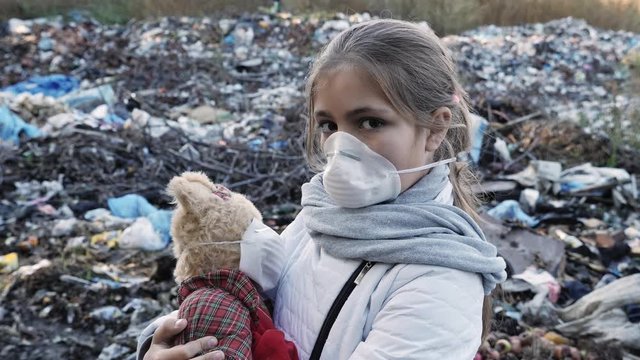 Young sad girl in a respirator on a dump with a plush toy looks into the camera. Ecology concept.