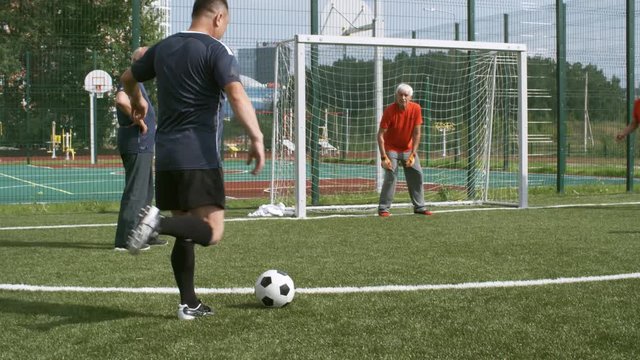 Mature man in sports clothing kicking football towards goal net, senior goalkeeper trying to catch it