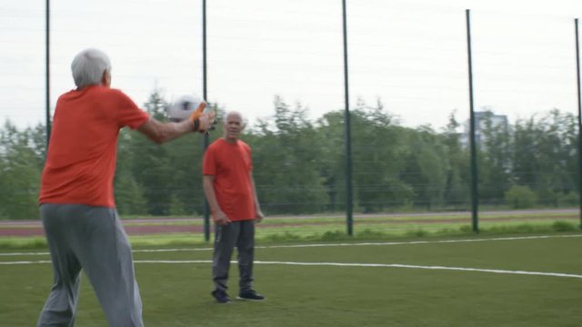 Mature man in sports clothes kicking football towards goal net, goalkeeper catching it and throwing back while teammates are watching