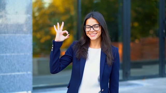 Successful pretty woman look to camera and showing OK sign on office building background. Professional female manager wearing glasses and suit. Student girl