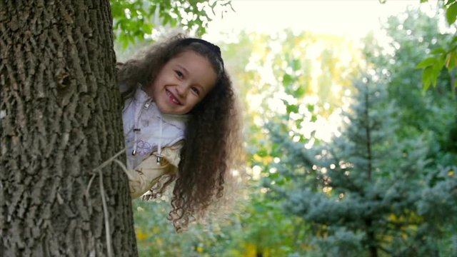 Beautiful Fashionable Happy Smiling Stylish Joyful European Little Cute Girl in a White Vest and Long Blond Curly Hair Walks through the Autumn Park, Looks out From Behind the Tree and Looks into the