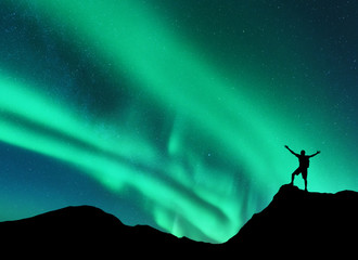 Northern lights and silhouette of standing man with raised up arms on the mountain peak in Norway....