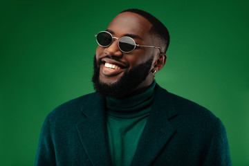 Fototapeta Man portrait. Style. Handsome Afro American guy in green jacket and sun glasses is looking at camera and smiling, on a green background obraz