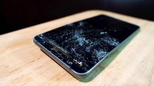 Smashed glass of mobile phone on wooden table rotating in light with half the screen broken and the frame bent